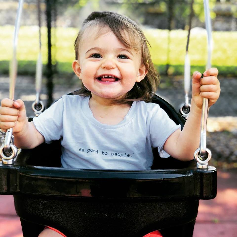 Baby wearing a Be Good To People shirt on the kids swingset