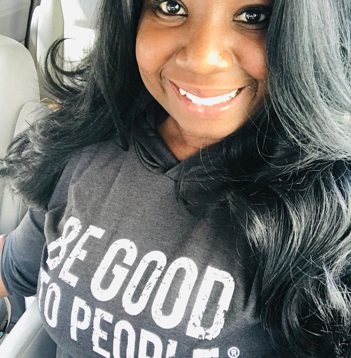 African-American woman with long hair and a beautiful smile wearing a hooded tee with Be Good to People on it in white. It's a close up photo and it looks like she is sitting in her car.
