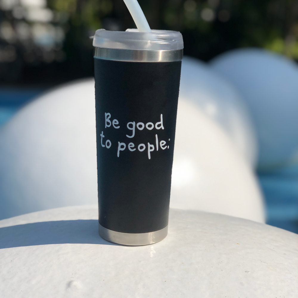A black travel coffee mug with Be Good to People writen in white resting on a large white ball. 
