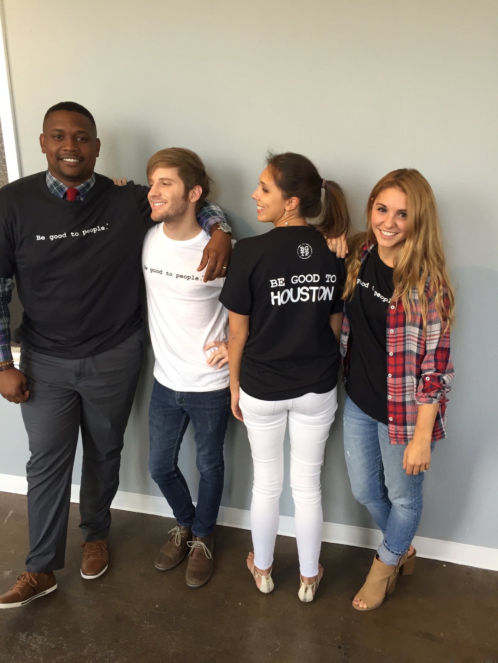 Four people standing against a wall, all wearing Be Good to People tshirts. One facing away with Be Good to Houston on the back of her shirt.. 