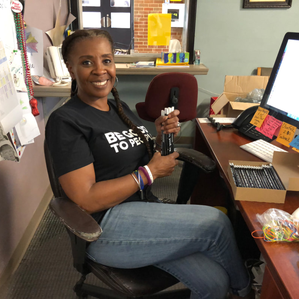 Woman at her desk wearing a Be Good to People tshirt and a stack of Be Good to People pencils in her hand and in a box on her desk