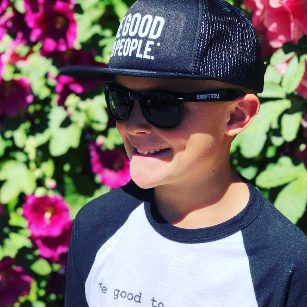 Young boy with a Be Good to People shirt and hat on and pink flowers in the background.